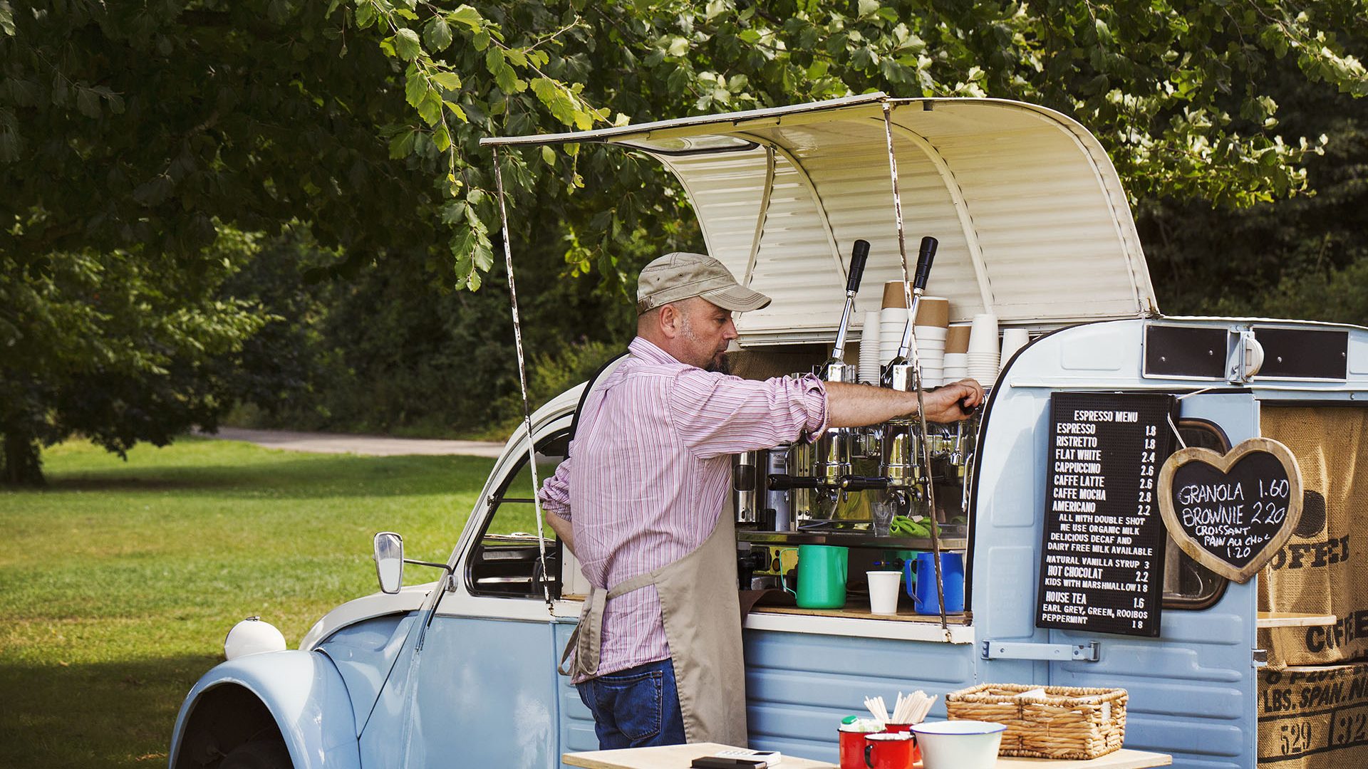 Bearded man wearing apron standing by blue mobile coffee shop, making coffee.